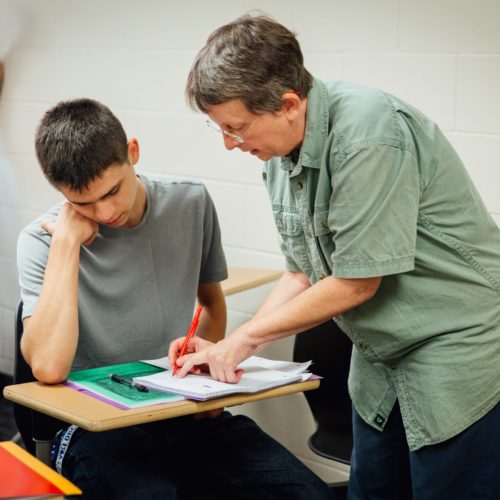 A teacher assists a student while another sits in the back with his hand raised