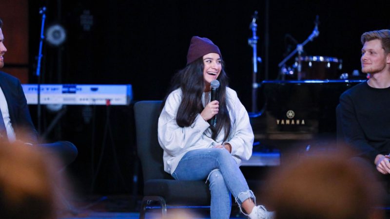 A woman with a microphone smiles while sitting on stage